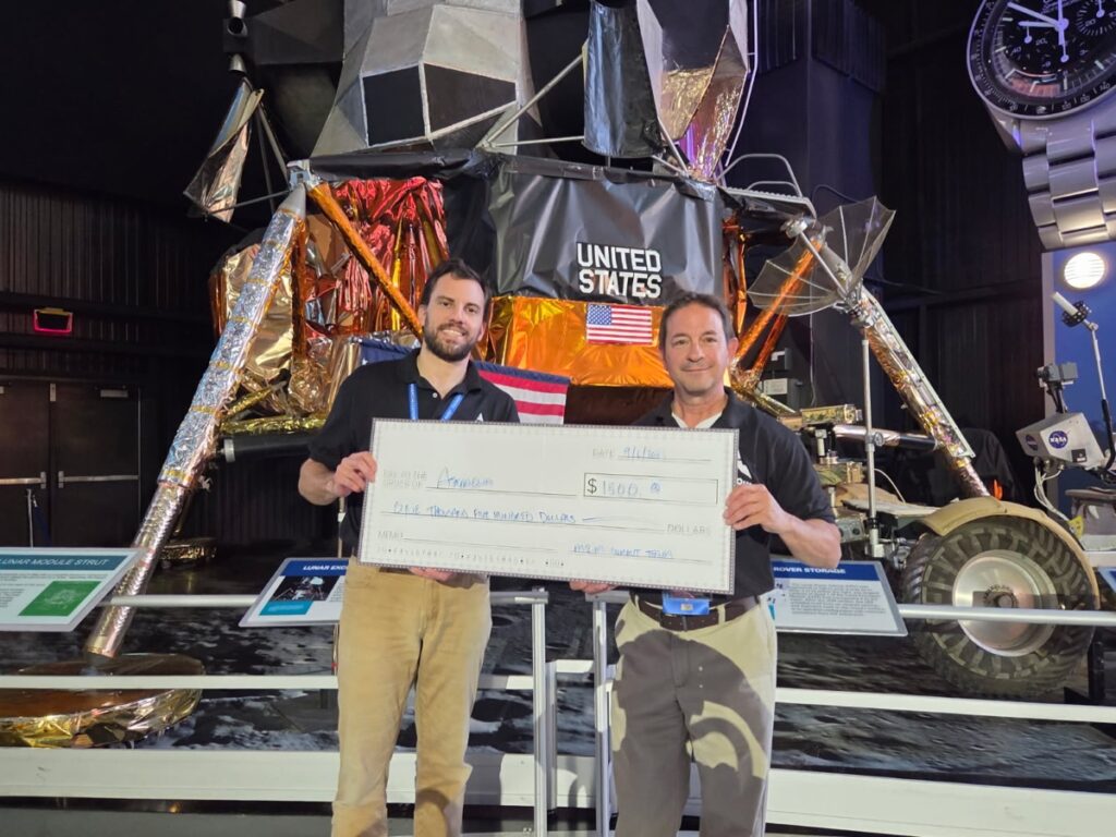 AstraQua representatives holding a second-place award check in front of a lunar module exhibit.
